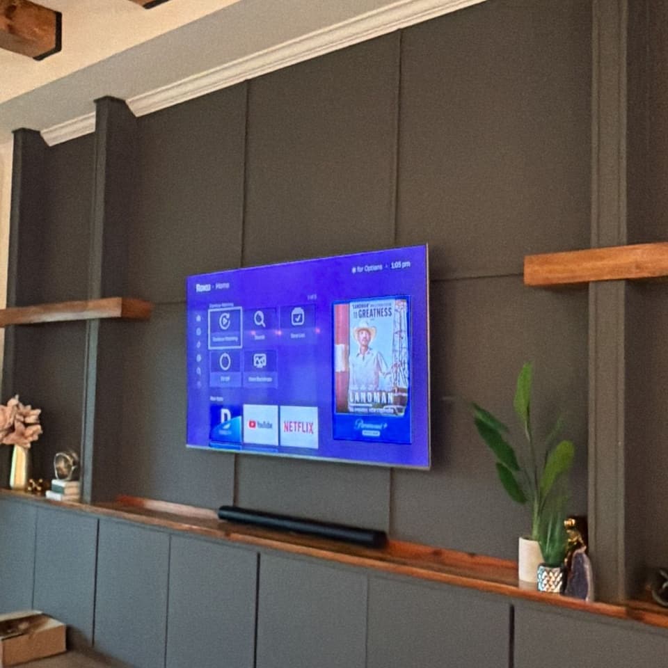 Televisor en una pared panelada color carbón bajo vigas de madera expuestas, con un estante flotante de madera y gabinetes oscuros debajo
