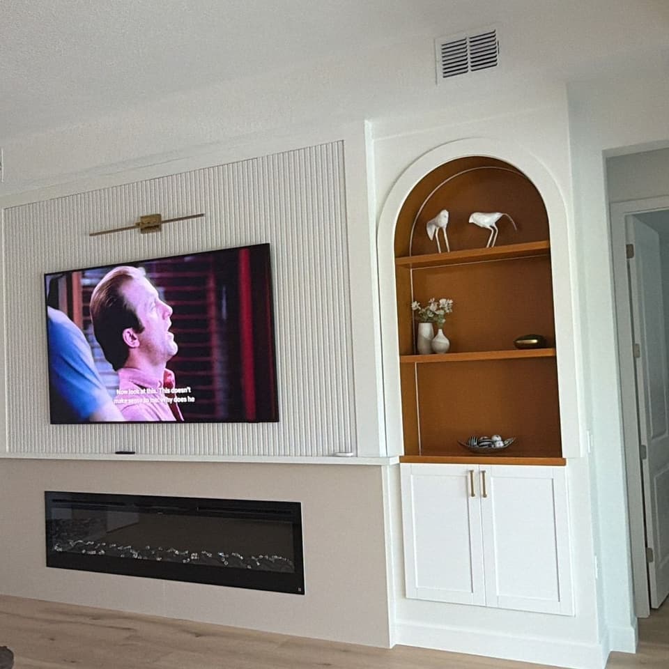 El mueble de pared terminado: dos nichos arqueados con repisas de madera, gabinetes bajos blancos tipo shaker, panel estriado detrás del televisor y chimenea lineal debajo