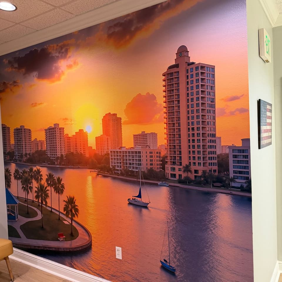 Gran mural fotográfico de un horizonte urbano frente al mar al atardecer, instalado de piso a techo en una sala interior
