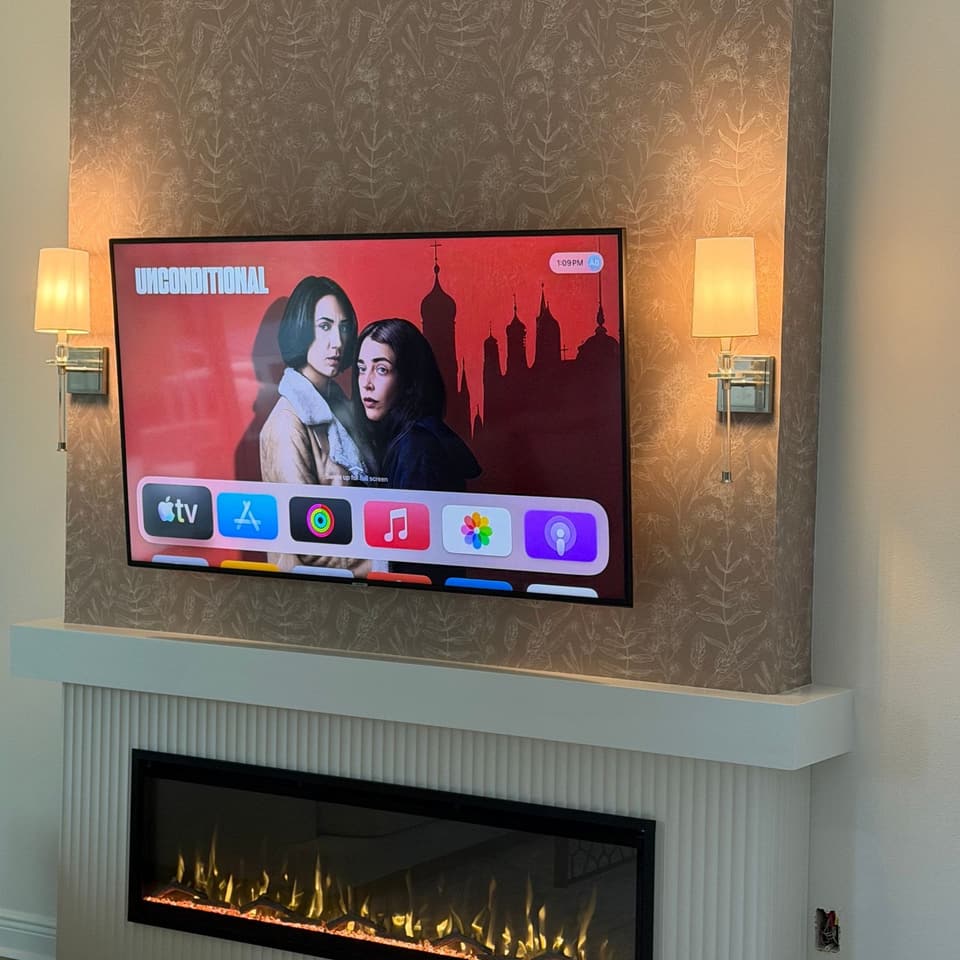 Televisor montado sobre una chimenea eléctrica lineal en una pared decorativa con papel tapiz, con enmarcado estriado y apliques de pared a los lados
