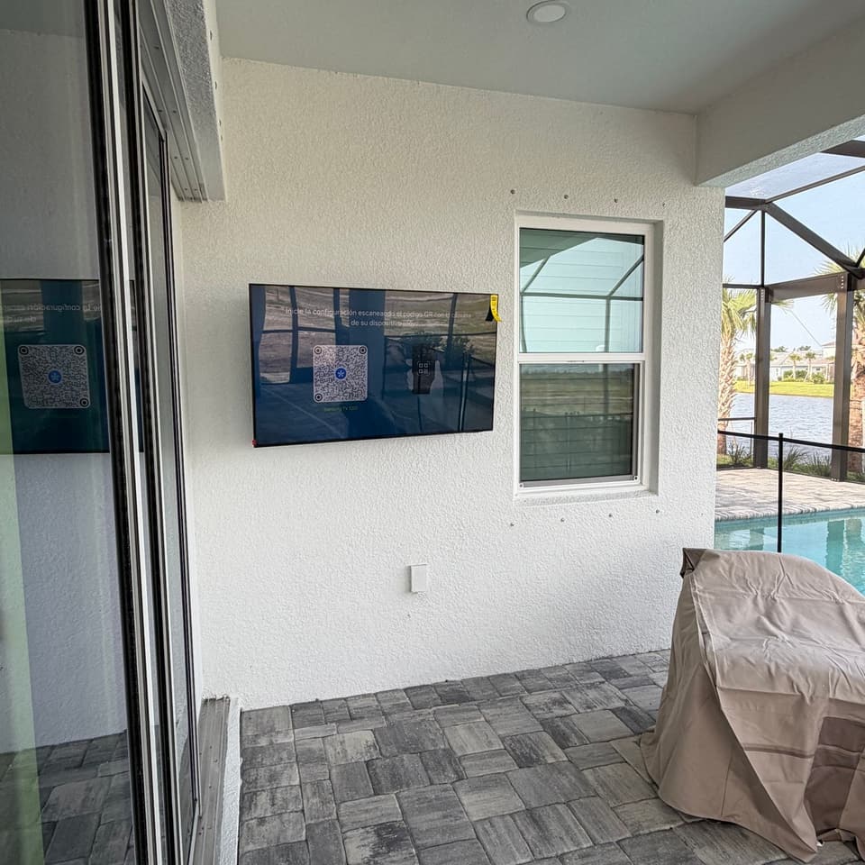Televisor montado en la pared exterior de una lanai con mosquitero, junto a una piscina y un patio de adoquines